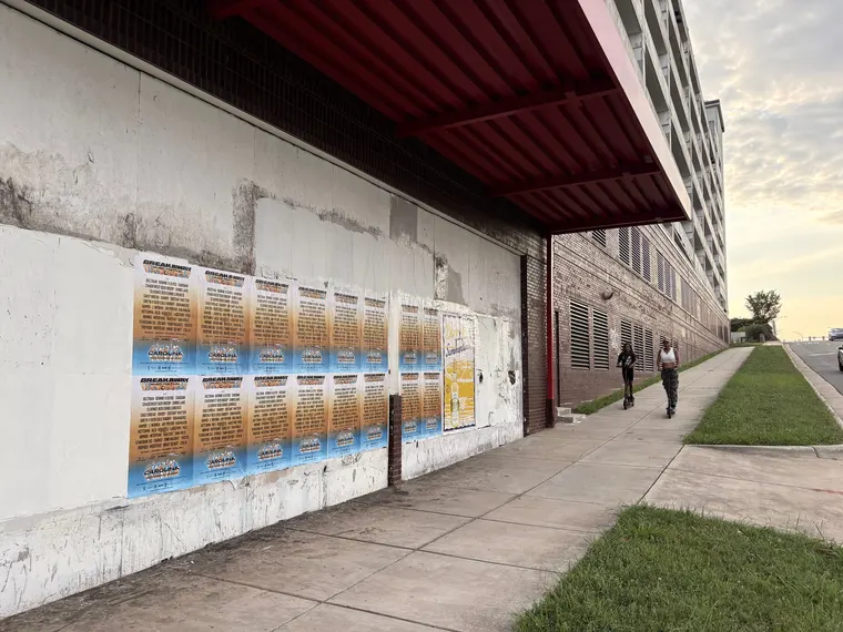 Breakaway Music Festival wheatpaste poster wall along a Charlotte sidewalk at golden hour, two people riding scooters past the paste-up, First Ward