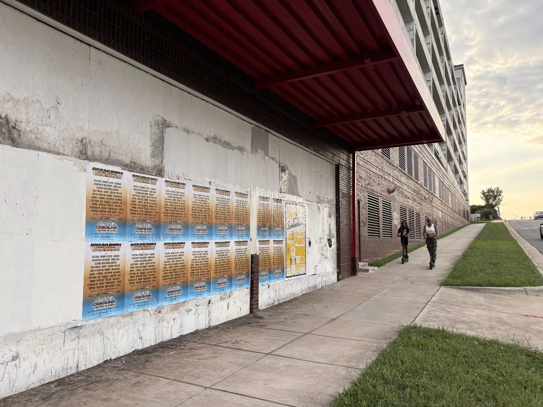 Breakaway Music Festival wheatpaste poster wall along a Charlotte sidewalk at golden hour, two people riding scooters past the paste-up, First Ward