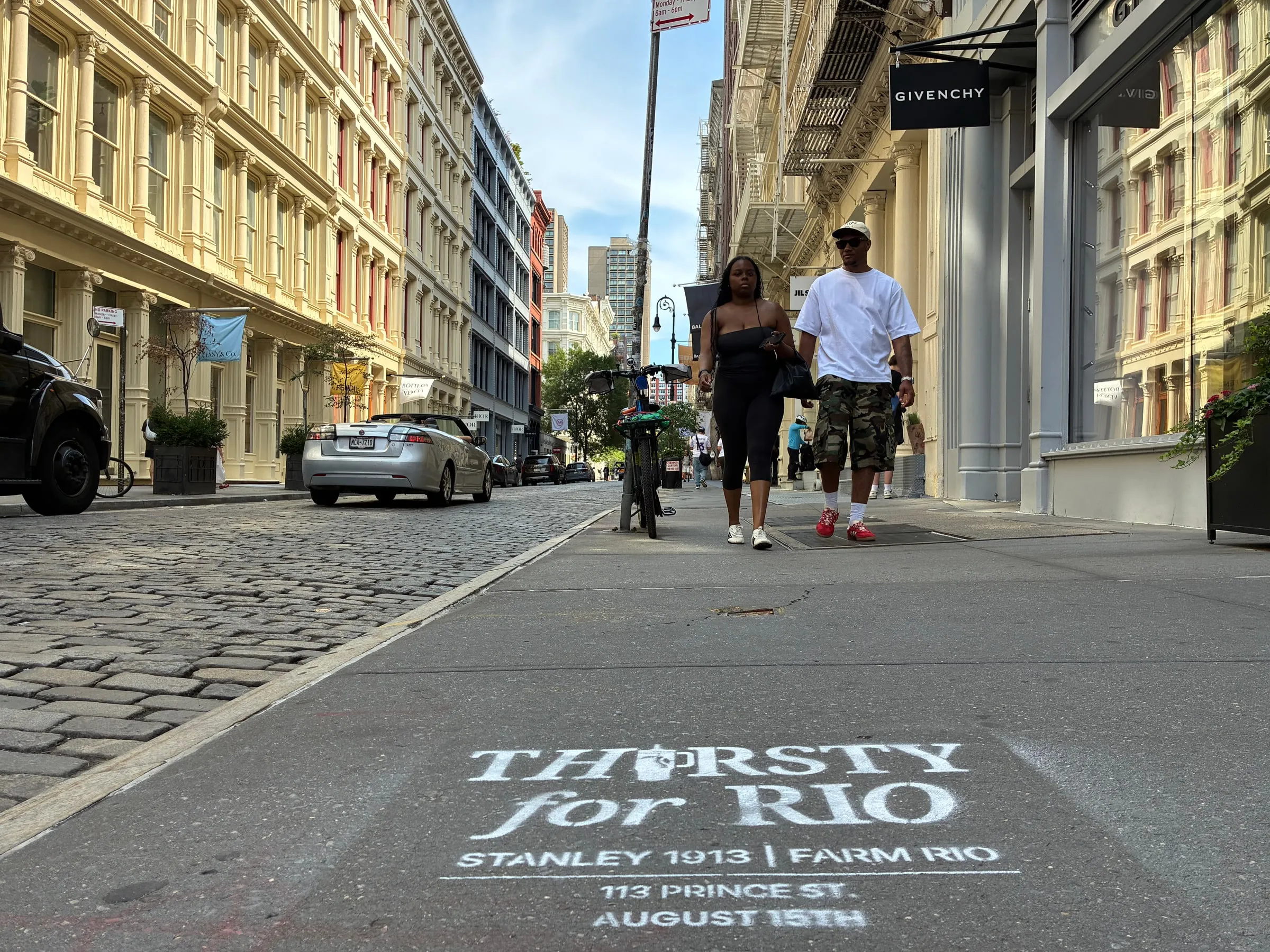 Pedestrians passing the stencil on a cobbled SoHo block with the Givenchy storefront beyond.