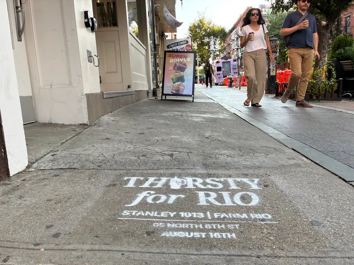 Farm Rio x Stanley 1913 chalk stencil reading Thirsty for Rio on the North 6th St sidewalk in Williamsburg, pedestrians and cafe signage beyond.