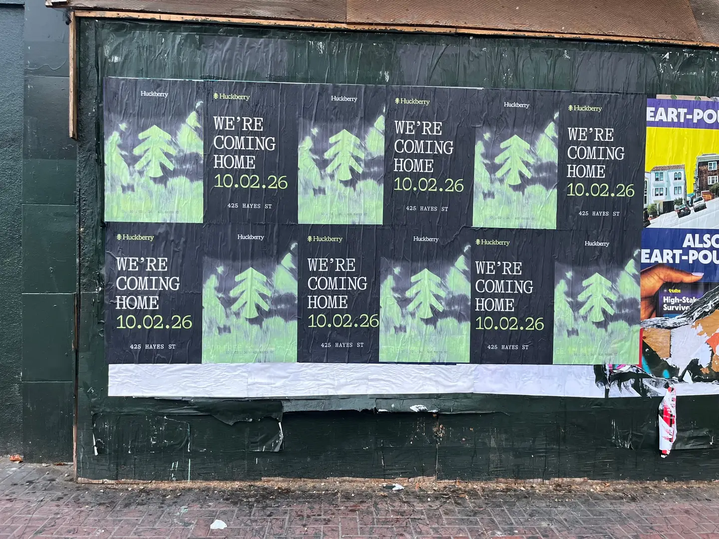 Huckberry wheatpaste posters close up on Market Street between Gough and Octavia: the type sheet reads We're coming home, 10.02.26, 425 Hayes St, between pine-tree illustrations