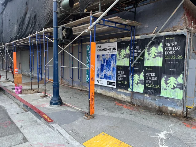 Huckberry wheatpaste posters on boards behind blue and orange scaffolding at Oak Street and Octavia Boulevard, Hayes Valley, beside an exhibition poster