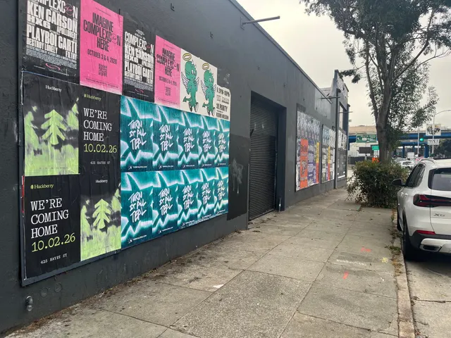 Huckberry wheatpaste posters among other event posters on a black wall on Bryant Street near 10th, SoMa, looking down the block