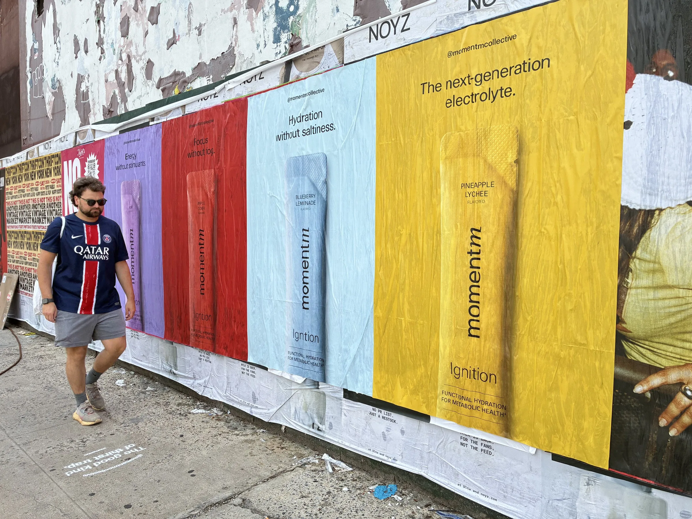 Angled Momentm poster run down a Williamsburg fence line with a pedestrian passing.