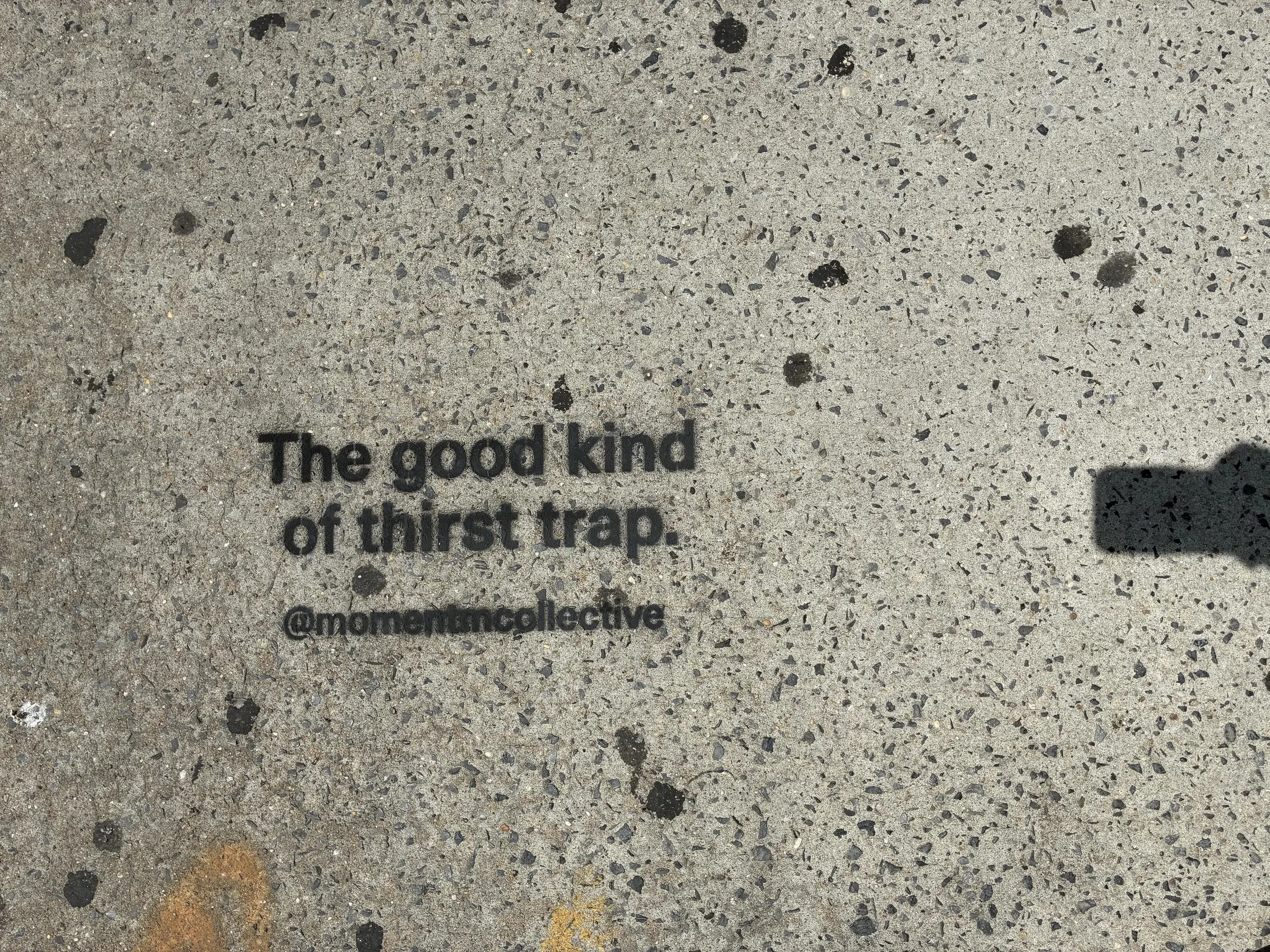 Momentm sidewalk stencil reading The good kind of thirst trap in black on light Williamsburg concrete.
