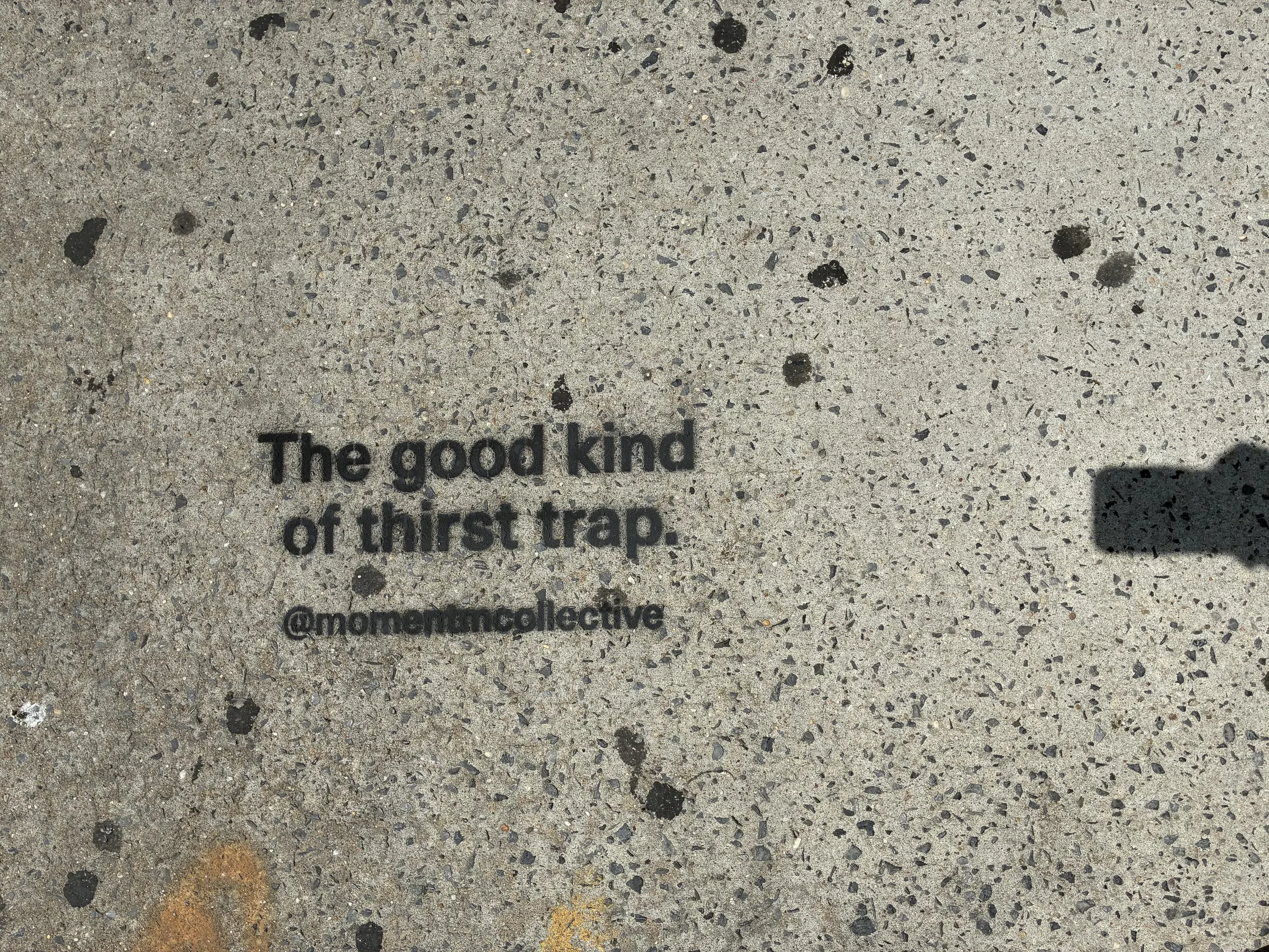Momentm sidewalk stencil reading The good kind of thirst trap in black on light Williamsburg concrete.