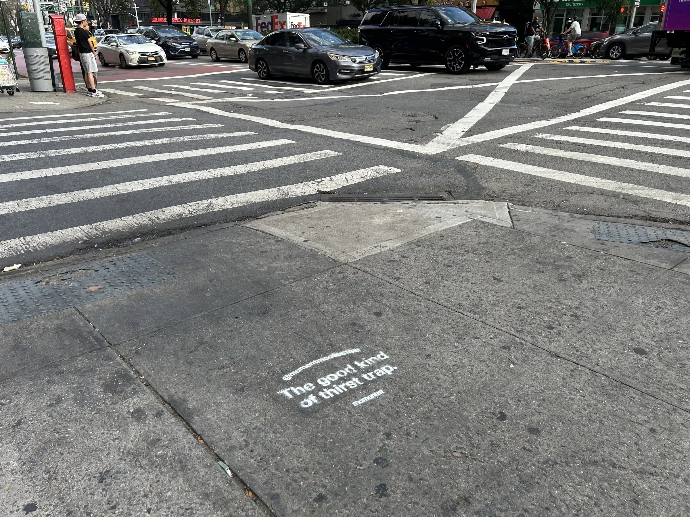 Momentm sidewalk stencil in white on dark pavement at a Lower East Side crosswalk in New York.