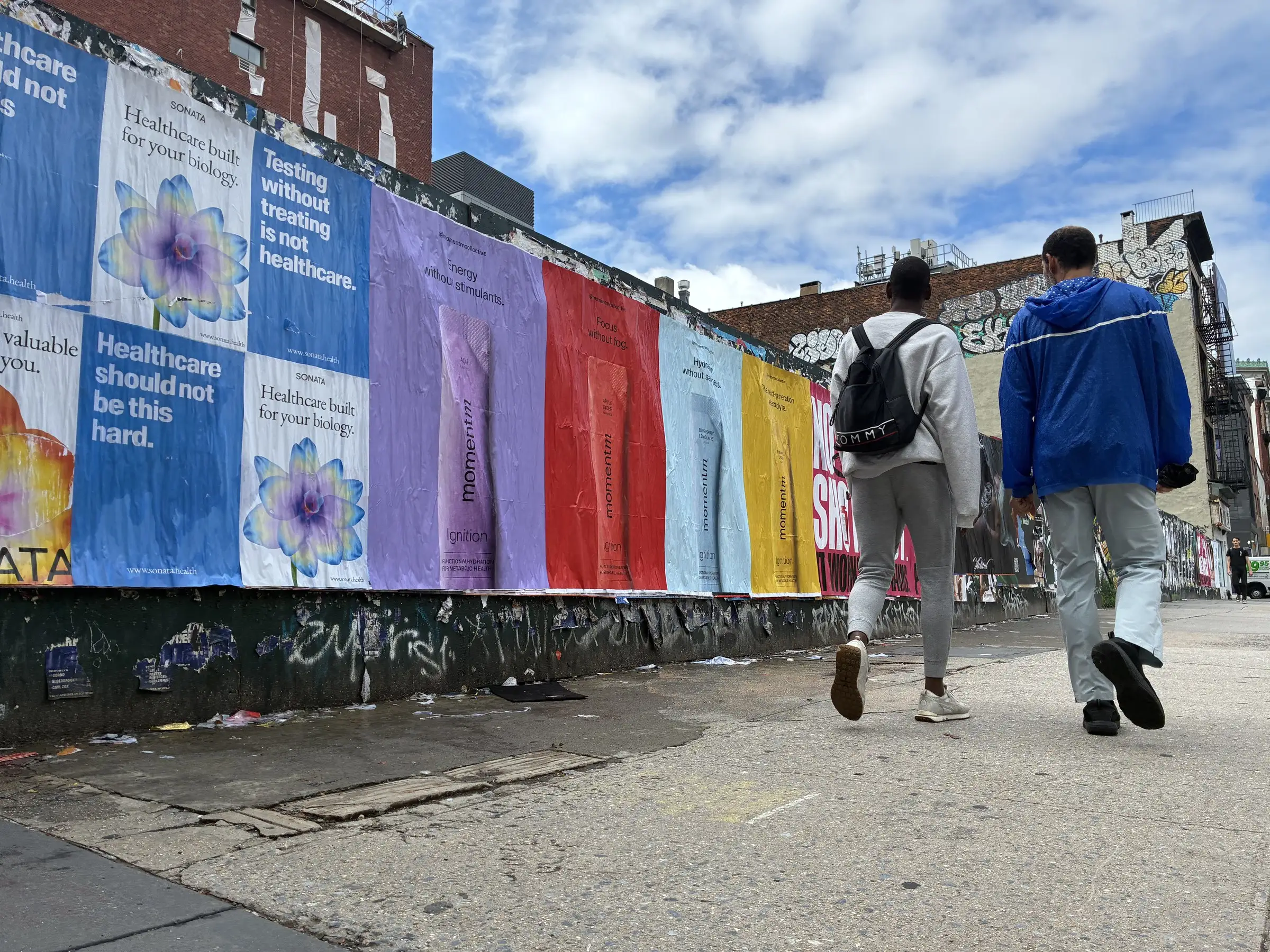 Pedestrians passing a run of Momentm jumbo 48x72 posters on Bowery-area construction hoarding in New York by Beyond Street Media.