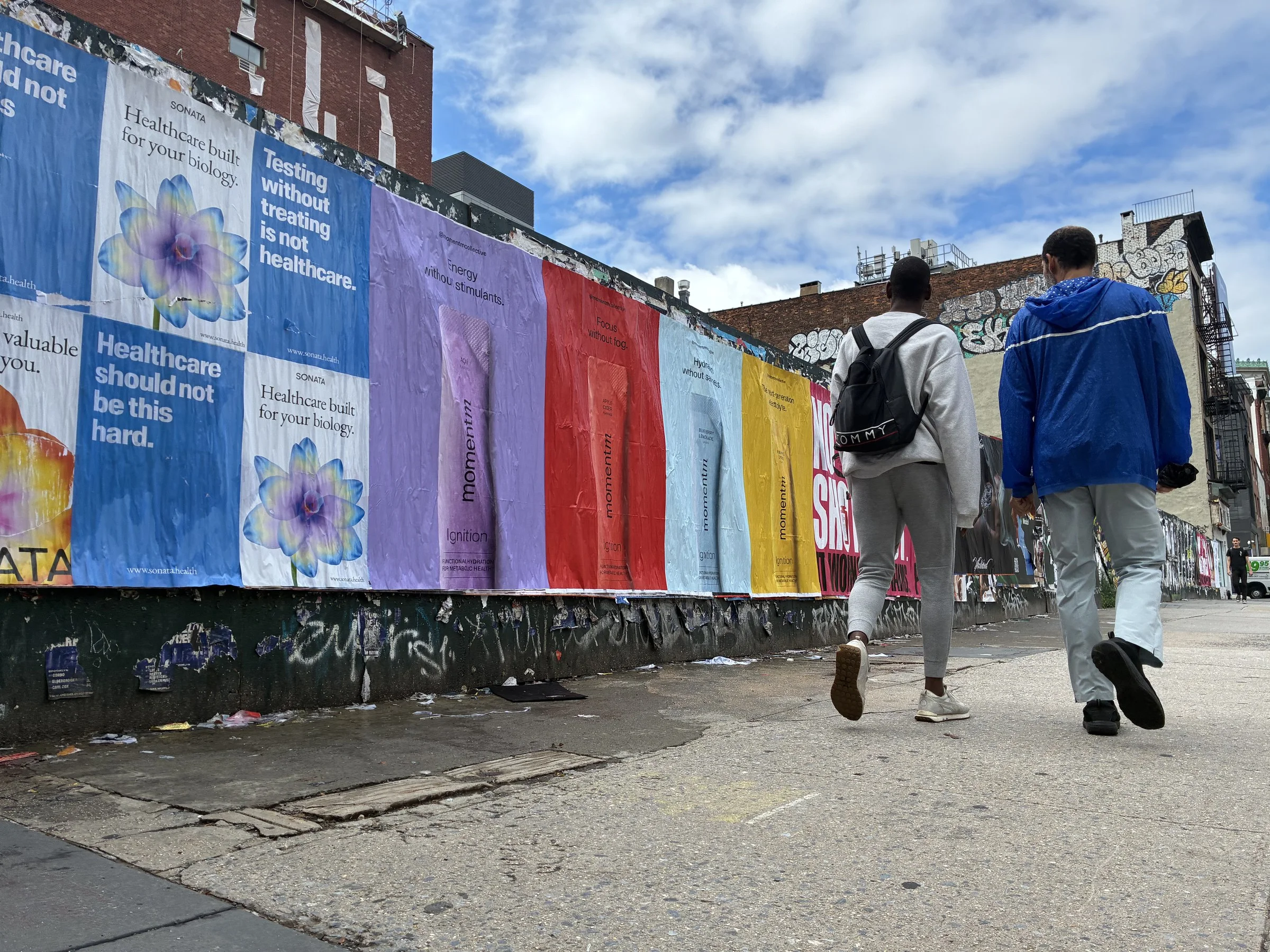 Pedestrians passing a run of Momentm jumbo 48x72 posters on Bowery-area construction hoarding in New York by Beyond Street Media.