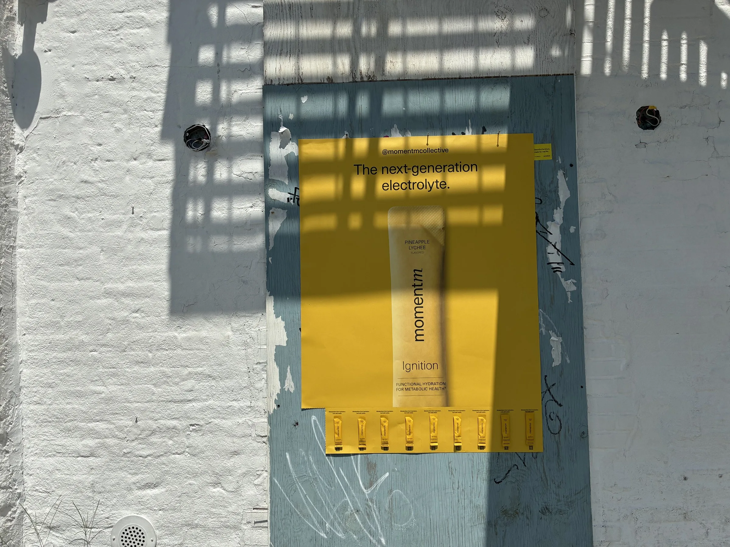 Momentm takeaway poster in yellow with a tear-off strip of product stick packs on a white brick Williamsburg wall.