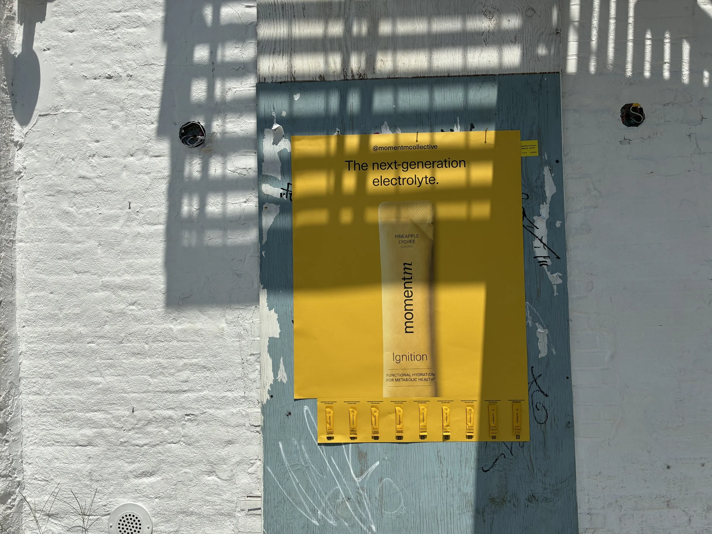 Momentm takeaway poster in yellow with a tear-off strip of product stick packs on a white brick Williamsburg wall.