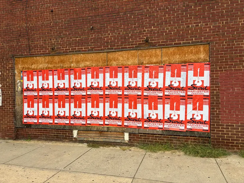 Scout Harvester wheatpaste in the red-orange colorway on brick above a boarded storefront at Charlotte Rail Trail, South End