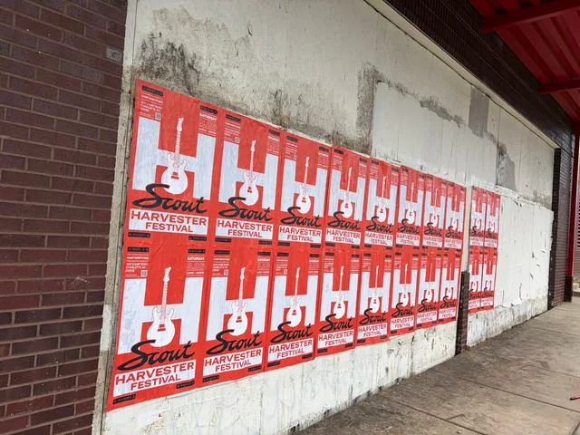 Scout Harvester wheatpaste in the red-orange colorway on a grimy white wall under a red steel eave, East 11th Street, First Ward