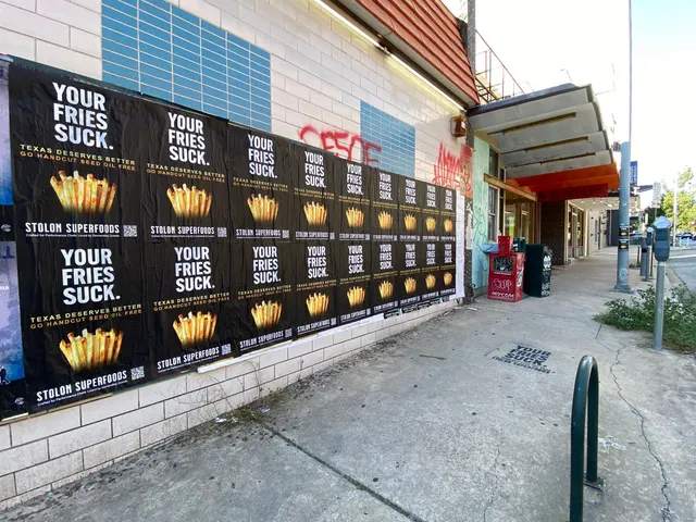Stolon Superfoods wheatpaste posters down a glazed-block wall, a black chalk stencil and parking meters ahead, on Lavaca Street at 16th, in daylight