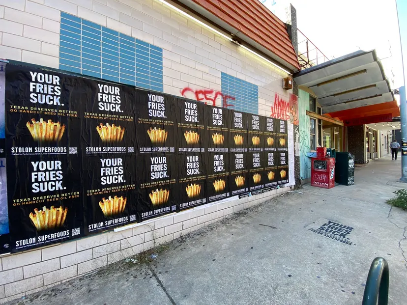 Stolon Superfoods wheatpaste posters, a two-row run under an orange tile roof, a black chalk stencil on the sidewalk, on Lavaca Street, in daylight