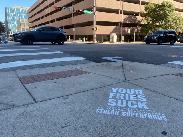 Stolon Superfoods chalk stencil in white at a crosswalk on Lavaca Street, the Lavaca blade and a parking garage behind, in daylight