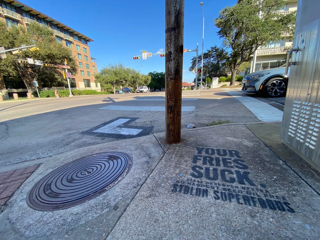 Stolon Superfoods chalk stencil in black beside a rusted pole and a manhole cover on Martin Luther King Jr Boulevard by the UT campus, in daylight