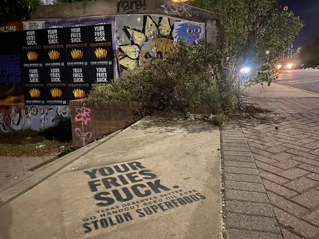 Stolon Superfoods chalk stencil in black below eight posters, four across in two rows, on a graffiti wall on South Congress Avenue, at night