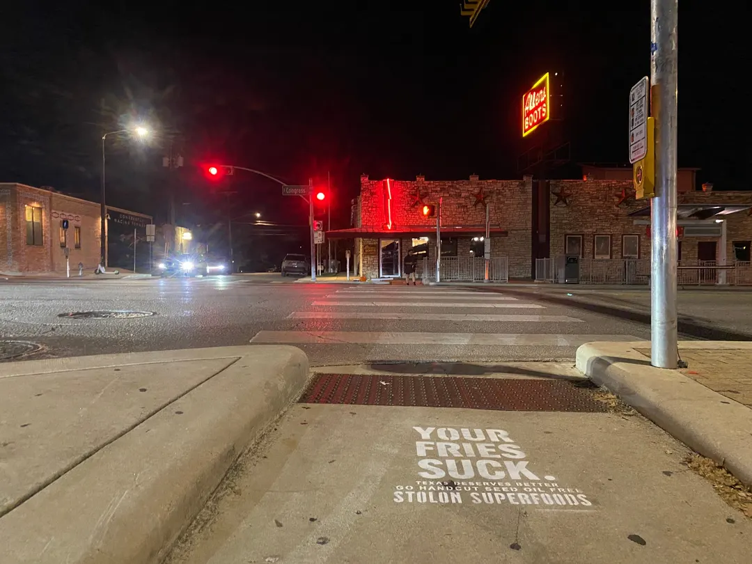 Stolon Superfoods chalk stencil in white on a crosswalk curb ramp on South Congress Avenue, at night, a neon BOOTS sign above a stone building