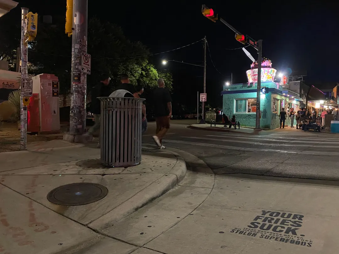 Stolon Superfoods chalk stencil in black on a curb corner on South Congress Avenue, at night, a giant lit ice cream cup sign across the street