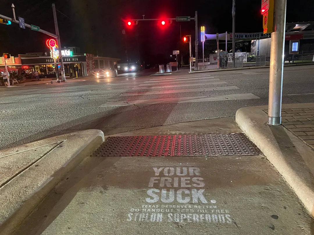 Stolon Superfoods chalk stencil in white on a curb ramp behind a tactile pad on South Congress Avenue, Congress and Elizabeth blades beyond, at night