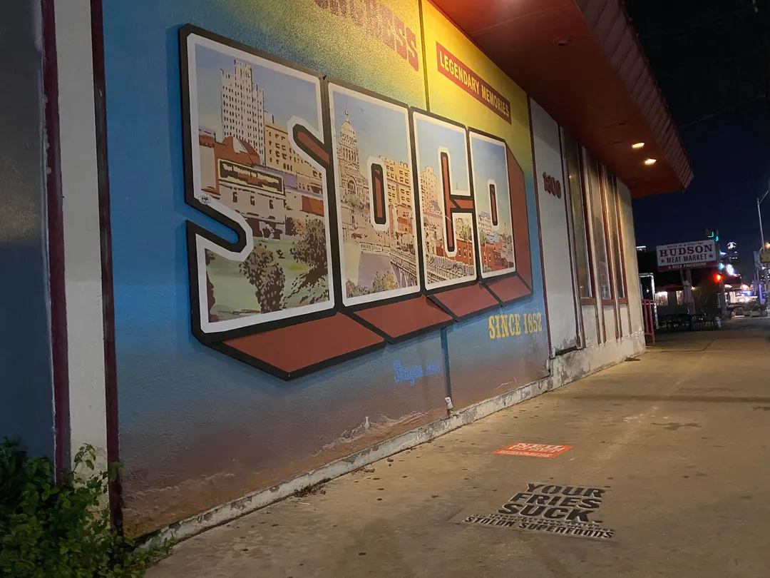 Stolon Superfoods chalk stencil in black on the sidewalk beside the SoCo postcard mural, Since 1852, on South Congress Avenue, at night