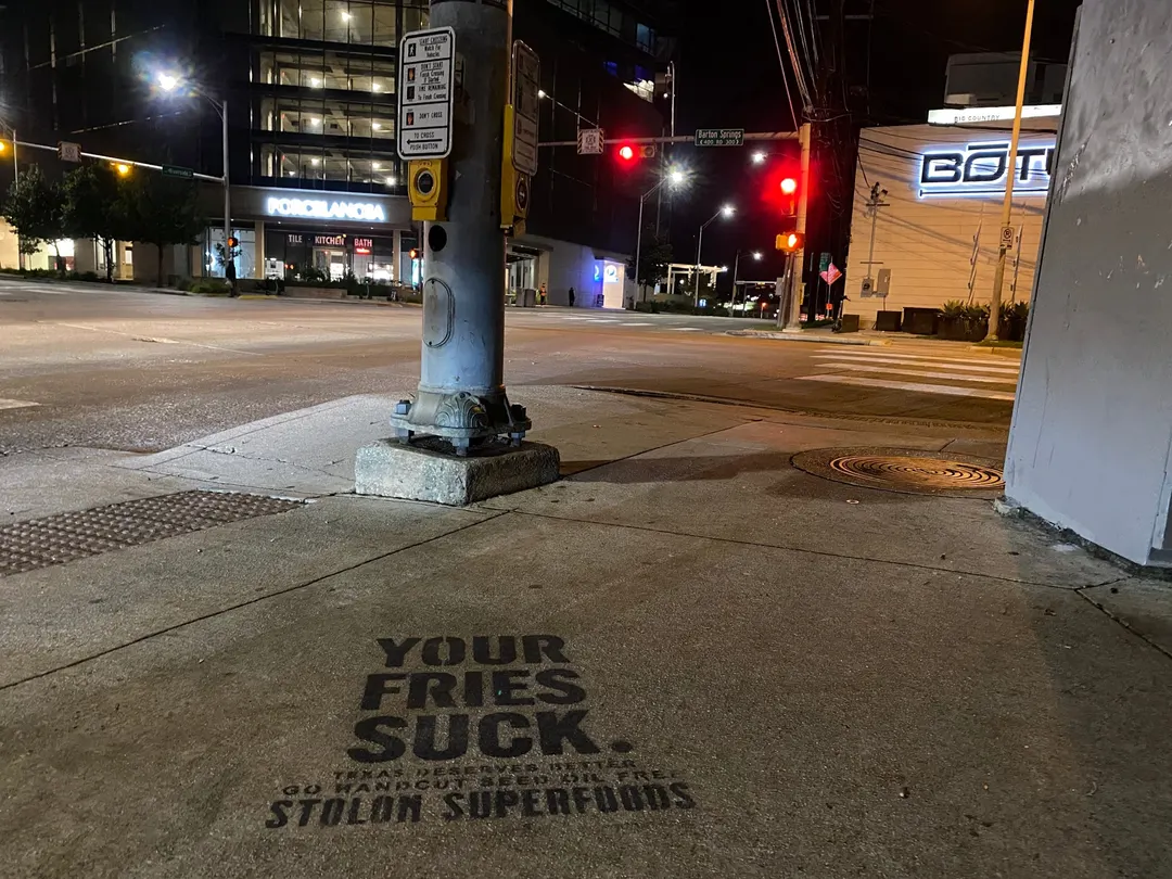 Stolon Superfoods chalk stencil in black in front of a crosswalk signal pole by Auditorium Shores, a Barton Springs street blade beyond, at night