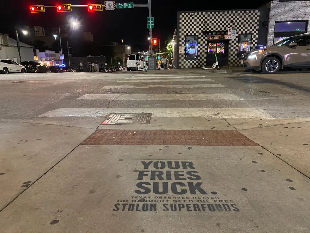 Stolon Superfoods chalk stencil in black on Red River Street at 7th, at night, facing a crosswalk and a checkerboard-tiled corner building