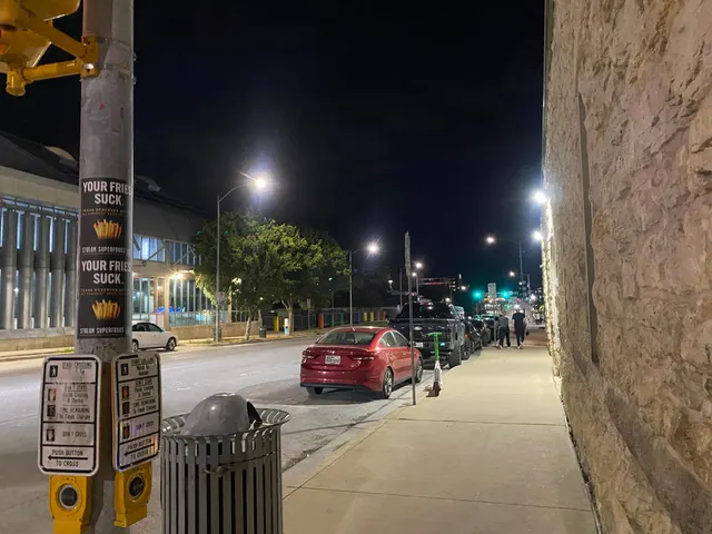 Stolon Superfoods pole stickers, two stacked over crosswalk placards on Red River Street at 7th, beside a rough limestone wall, at night
