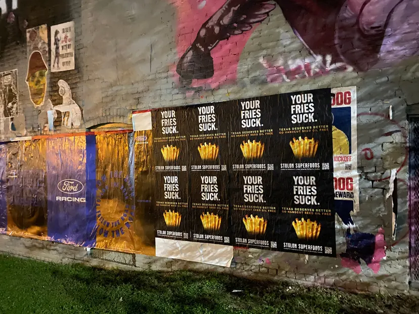 Stolon Superfoods wheatpaste posters, eight in two rows of four, under a pink mural on East 6th Street at San Marcos, East Austin, at night