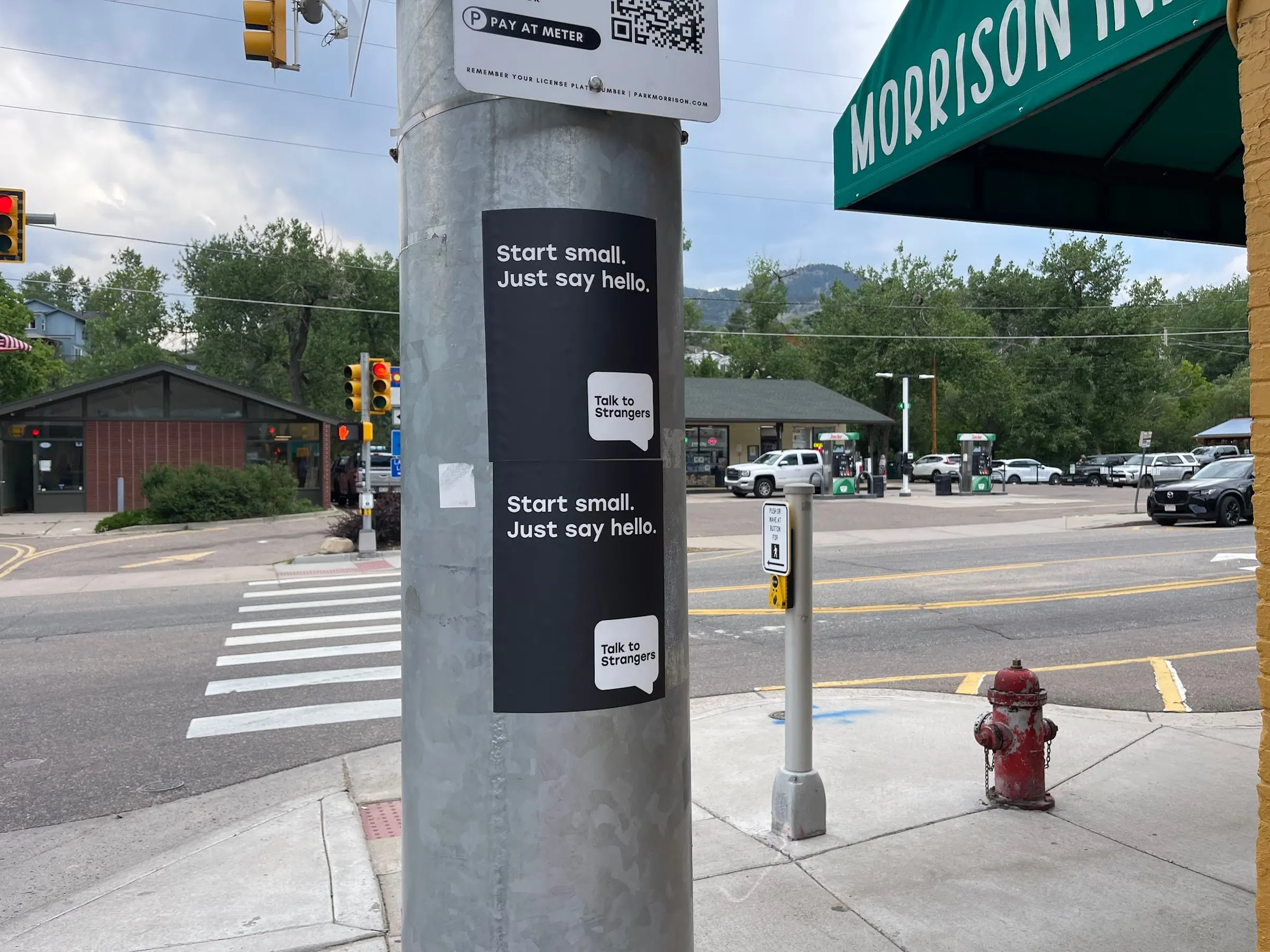 Talk to Strangers pole sticker install by Beyond Street Media at 301 Bear Creek Ave, Morrison, Colorado — two vertical 'Start small. Just say hello.' pole stickers at a crosswalk, foothills behind.