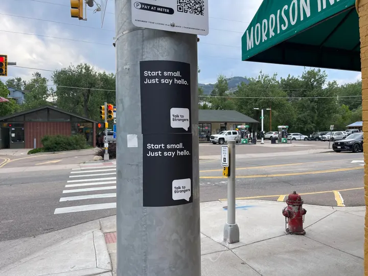 Talk to Strangers pole sticker install by Beyond Street Media at 301 Bear Creek Ave, Morrison, Colorado — two vertical 'Start small. Just say hello.' pole stickers at a crosswalk, foothills behind.