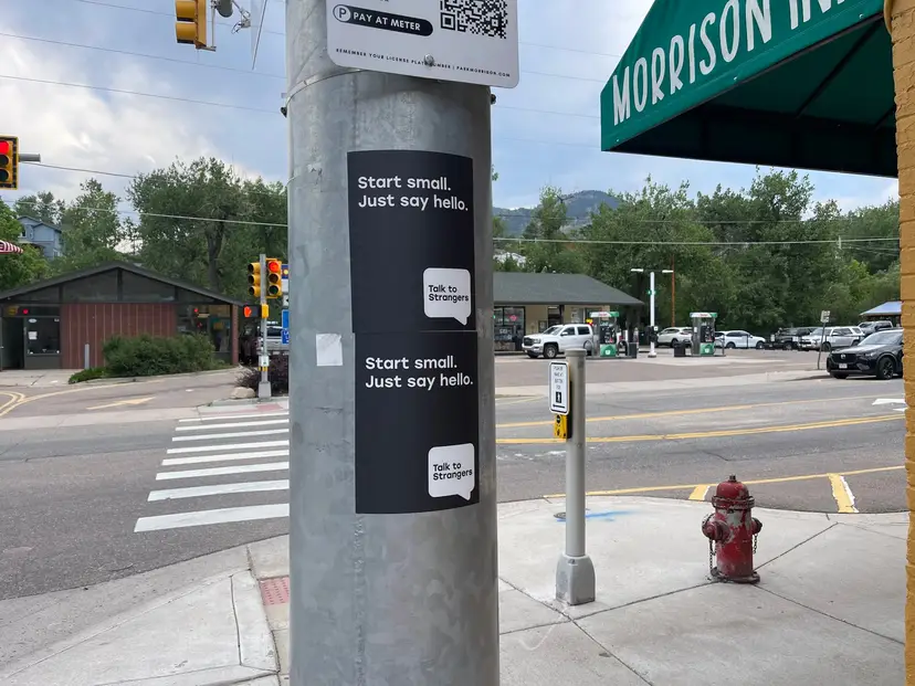 Talk to Strangers pole sticker install by Beyond Street Media at 301 Bear Creek Ave, Morrison, Colorado — two vertical 'Start small. Just say hello.' pole stickers at a crosswalk, foothills behind.