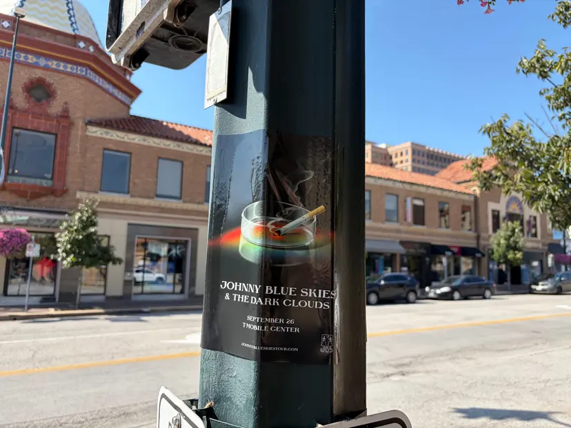 Johnny Blue Skies ashtray-design pole snipe at Country Club Plaza, poster below a pedestrian signal box, red sedan passing by