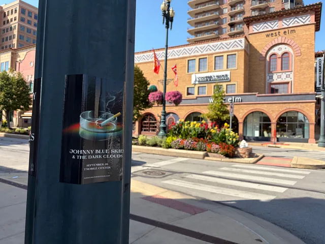 Johnny Blue Skies ashtray poster on a pole at Country Club Plaza, West Elm arched brick facade and flower beds across the crosswalk