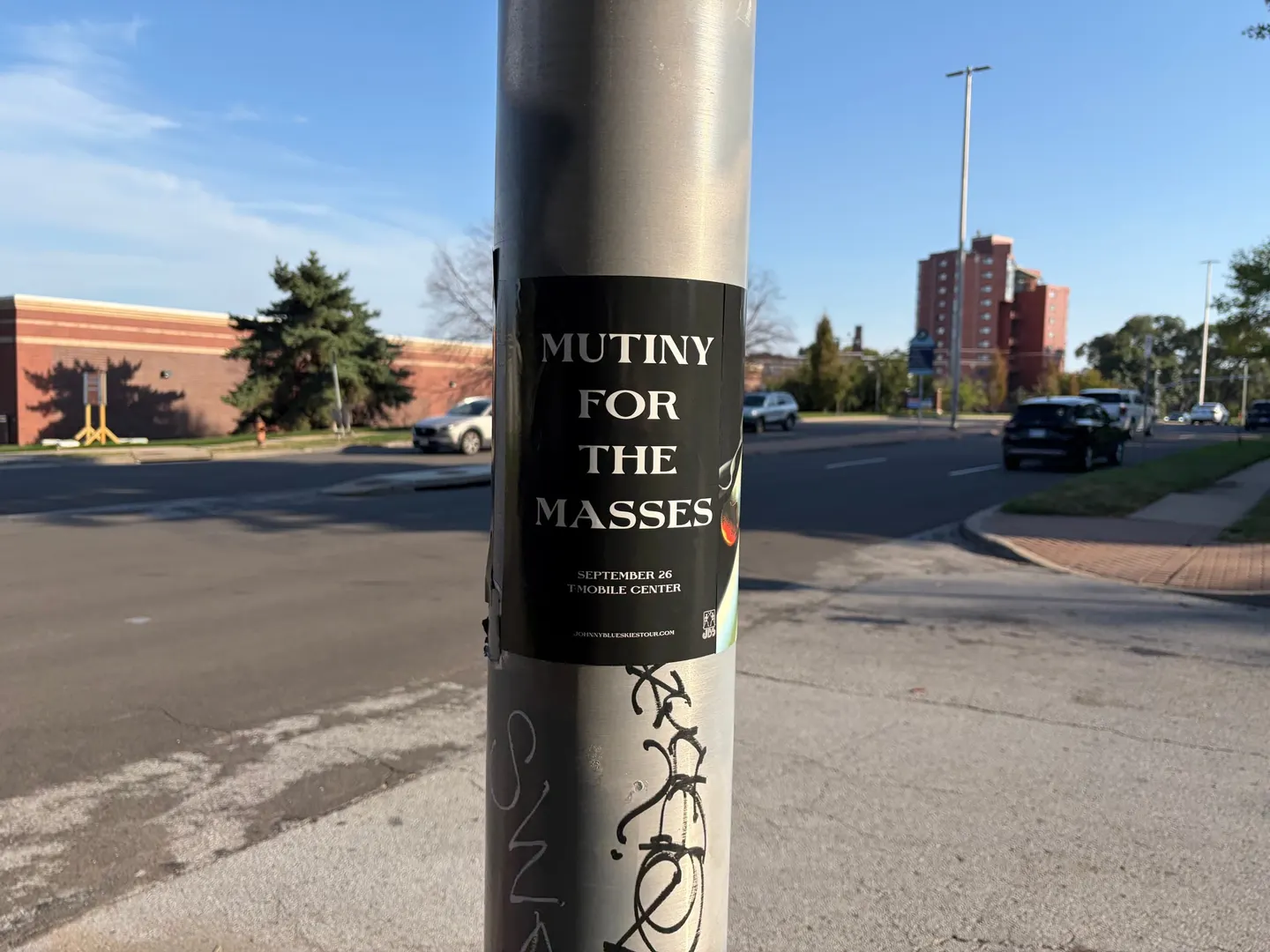 Mutiny for the Masses pole snipe on a brushed aluminum pole at Mill Street, Westport, graffiti tags below it and a brick building behind