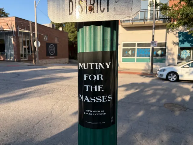 Mutiny for the Masses pole snipe below a District street sign on Broadway Boulevard, Westport, graffiti-tagged storefront glass behind it